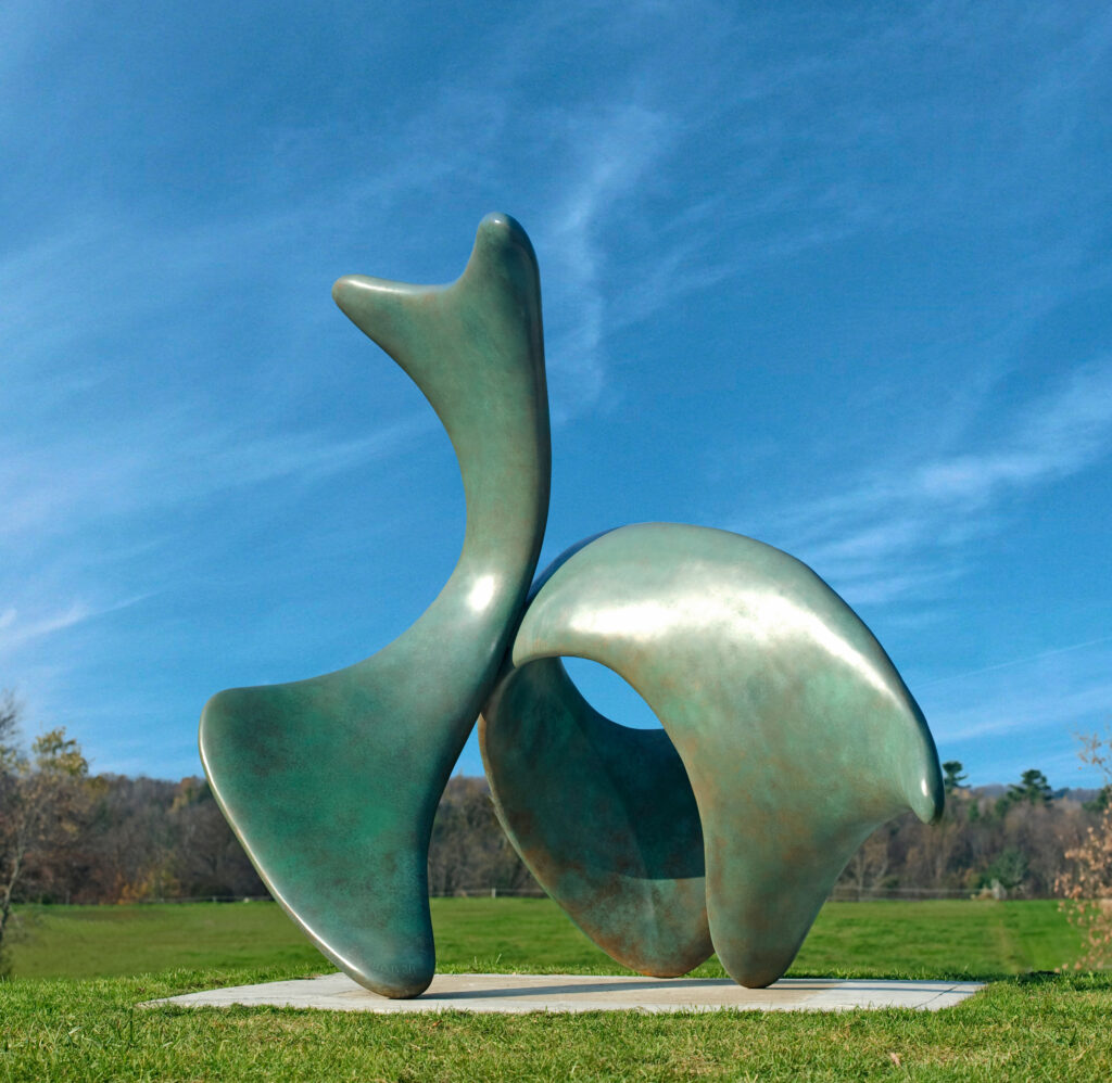 A large abstract bronze sculpture with a green patina sits on green grass in front of a blue sky