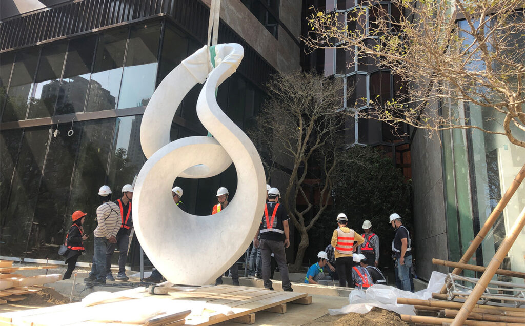 A monumental sculpture carved from Carrara marble hangs from a strap during installation on a city street. 