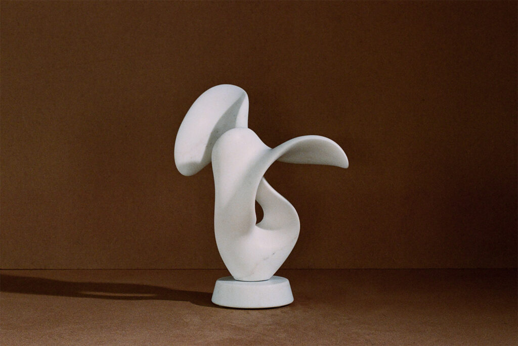 Muse, a white abstract sculpture carved from Carrara marble on a brown table. its flattened planes and curves turn in on itsself like a secret. 