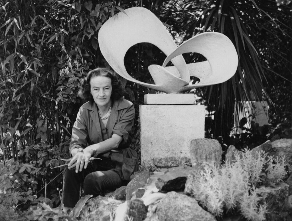 Sculptor Barbara Hepworth sits in a garden with an abstract sculpture