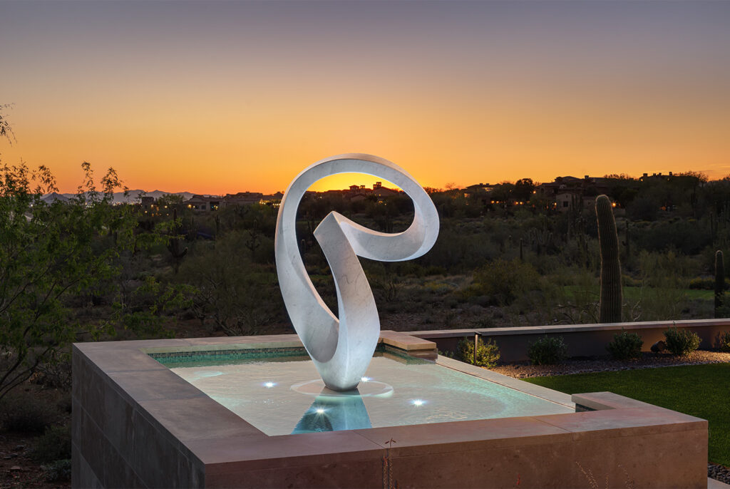 A looping abstract marble sculpture sits in a reflecting pool in front of a yellow and blue sunset